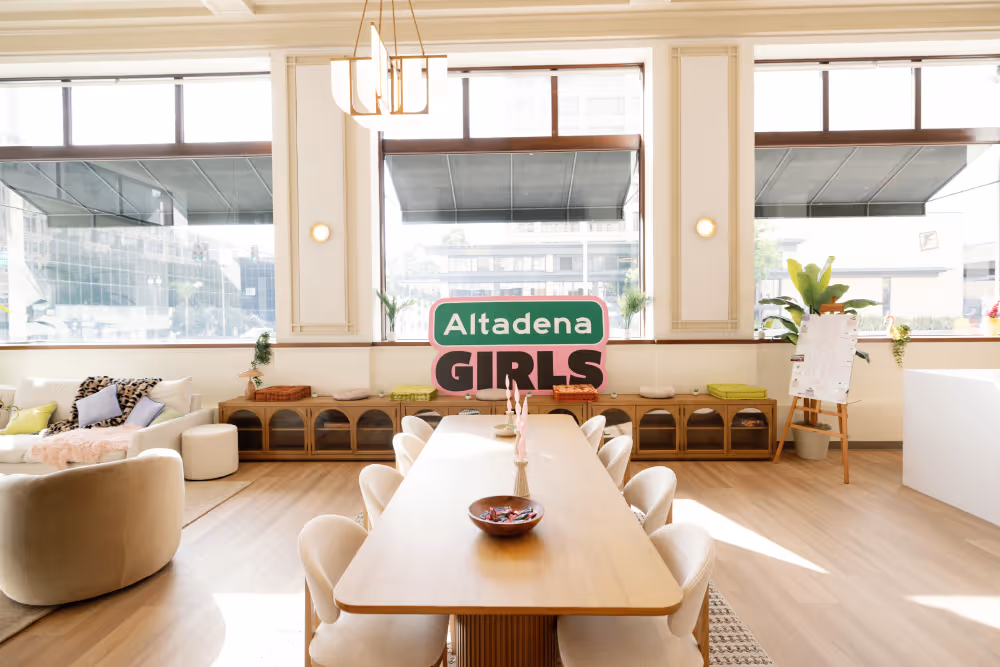 Bright modern meeting room with wooden table, white chairs, and large windows showing city view.