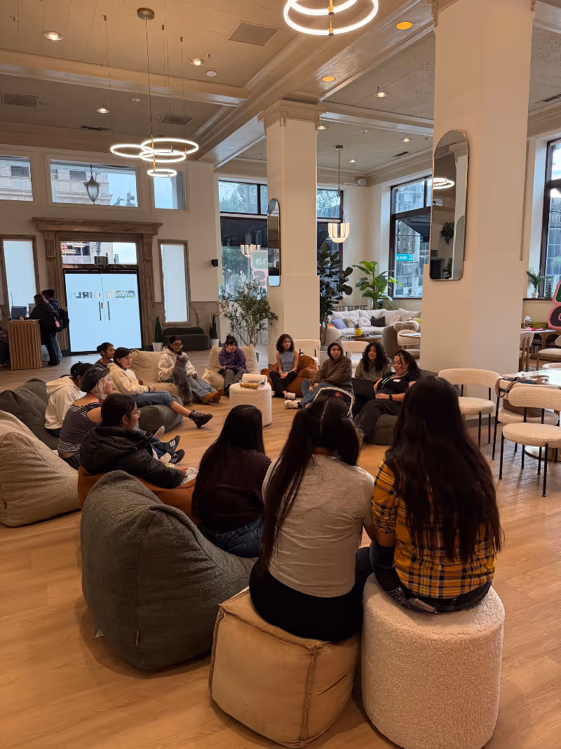 Group of people sitting in a circle in a modern, well-lit room with large windows and hanging lights.