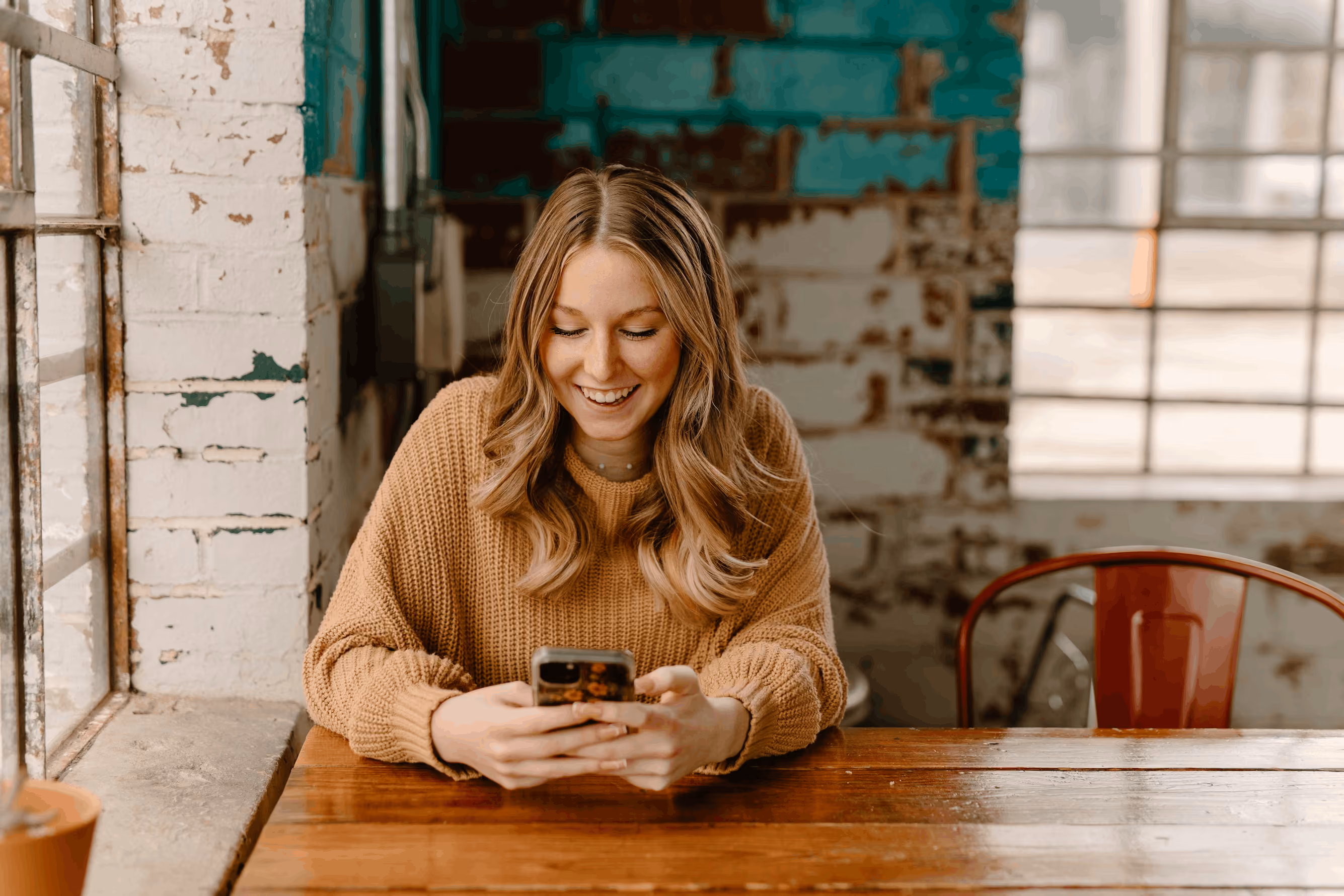Smiling woman in a brown sweater looking at her phone while sitting at a wooden table near a window.