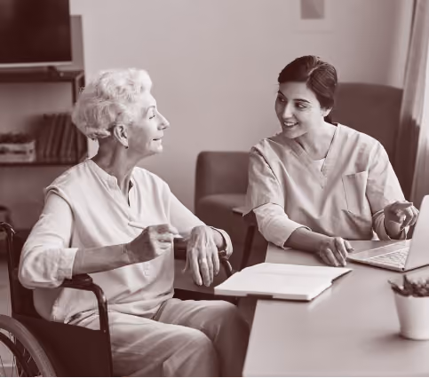 Healthcare worker in scrubs smiling and talking with elderly woman in a wheelchair at a table.