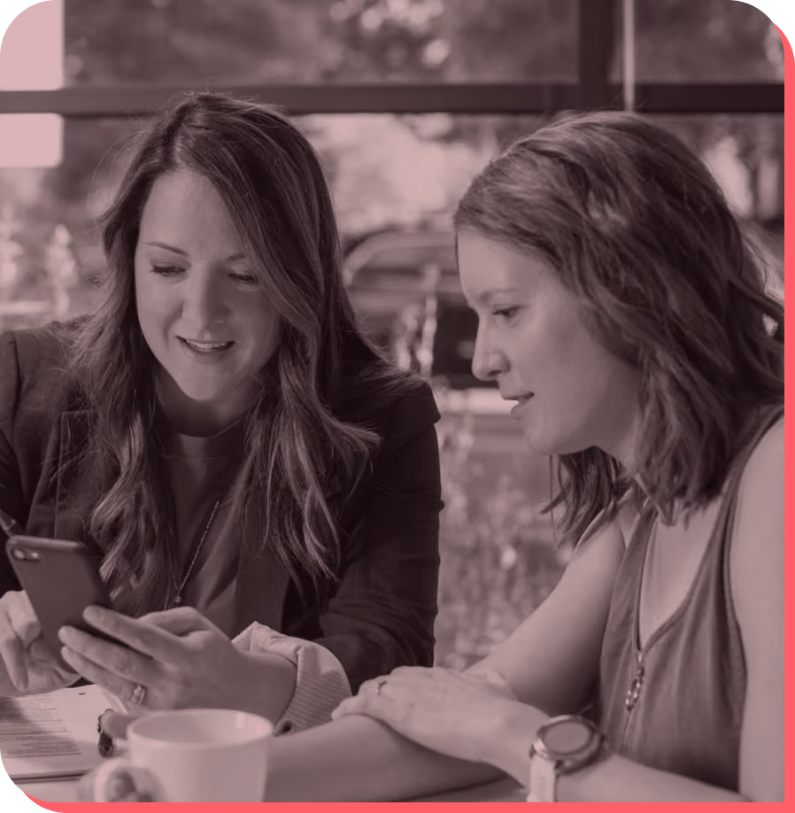 Two women sitting at a table, looking at a smartphone together with a coffee cup nearby.