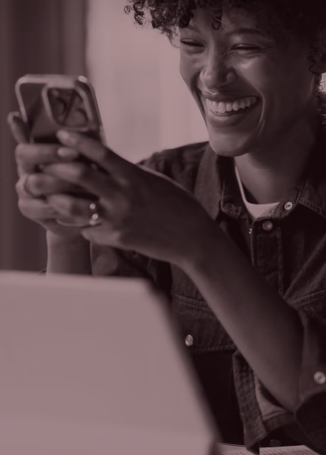 Smiling woman with curly hair holding a smartphone and looking at its screen, sitting at a desk.