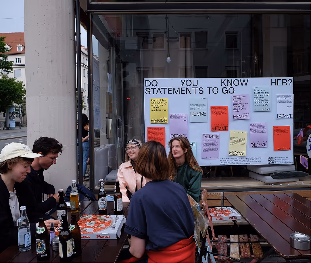 Group of five people sitting around an outdoor table with pizza and drinks, in front of a large window displaying a poster titled 'Do You Know Her? Statements to Go' with colorful notes.