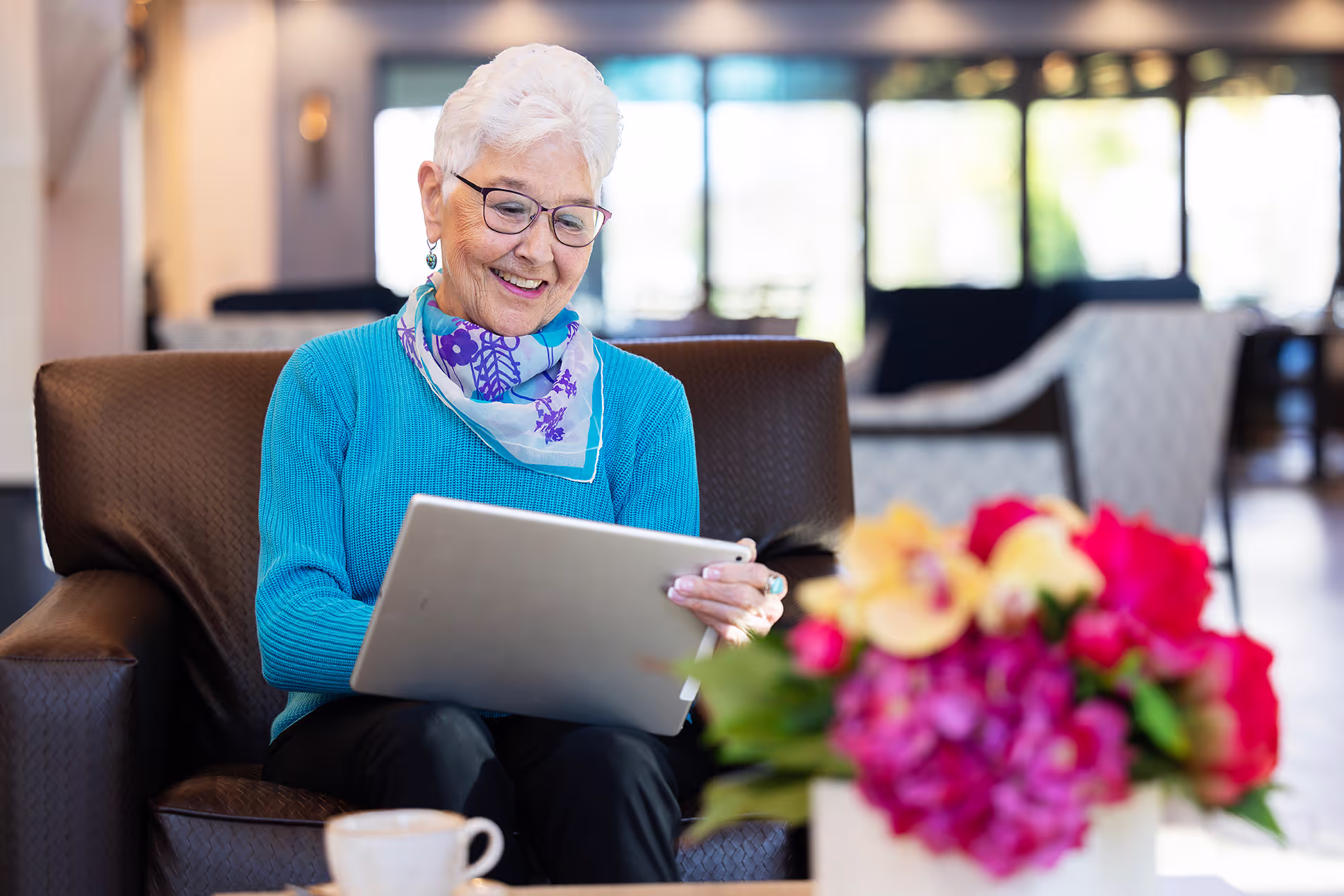 Senior woman using an iPad in a large common space, comfortably engaged in technology while surrounded by a spacious, inviting environment.