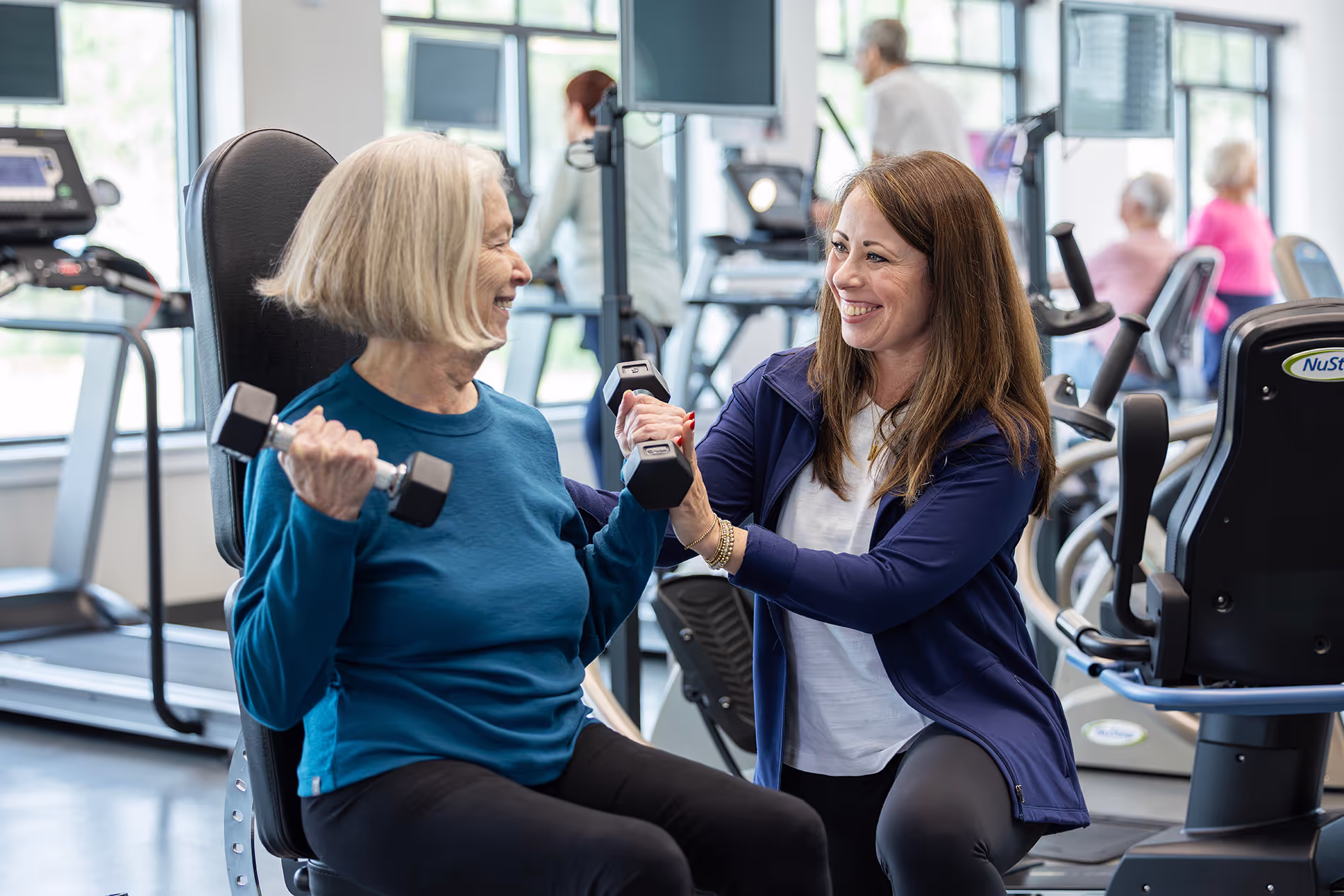 Trainer assisting a senior woman with lifting weights at the gym, providing guidance and encouragement in a supportive fitness environment.