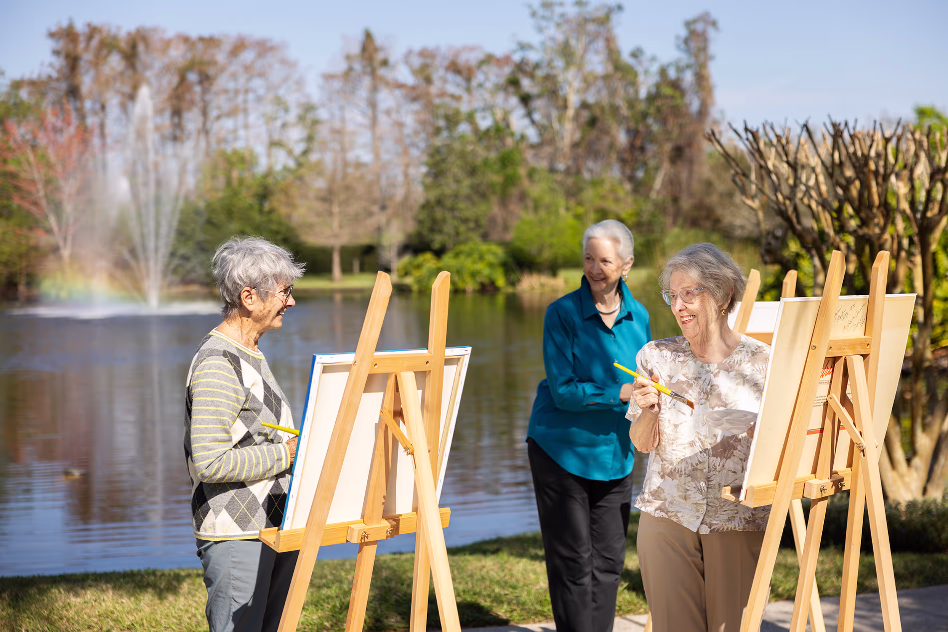 Group of older women painting on easels and canvases by the lake, enjoying a peaceful outdoor art session surrounded by nature.
