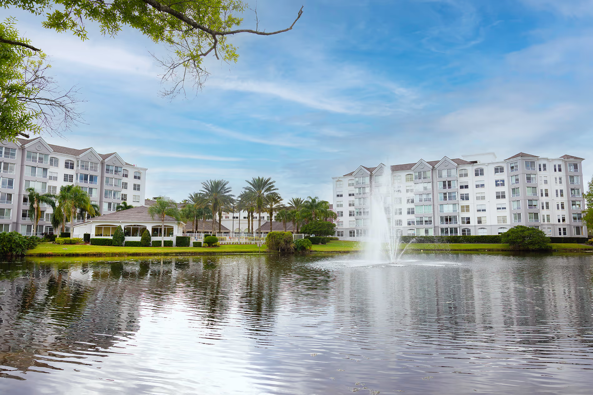Two apartment buildings standing behind a scenic lake with a central water feature, surrounded by landscaped greenery and a calm, inviting atmosphere.
