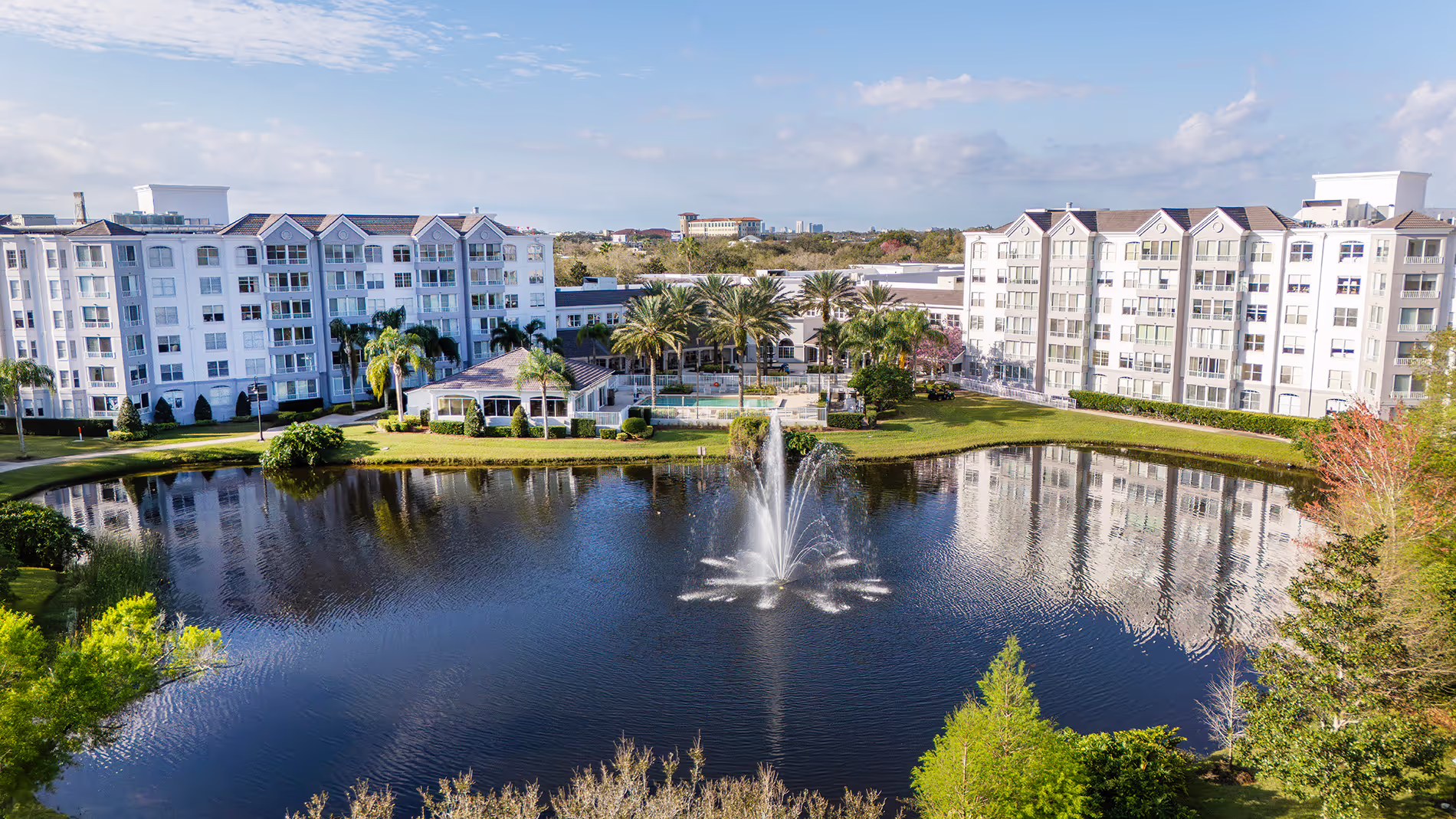Two apartment buildings positioned behind a tranquil lake, featuring a beautiful water fountain that adds to the peaceful ambiance of the scene.
