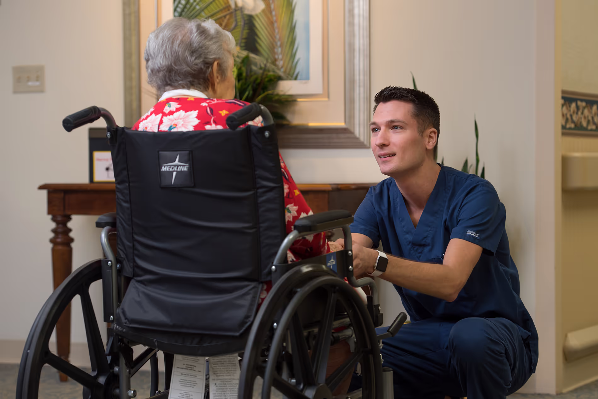 Male nurse leaning in front of a resident in a wheelchair, offering assistance and providing attentive, caring support.