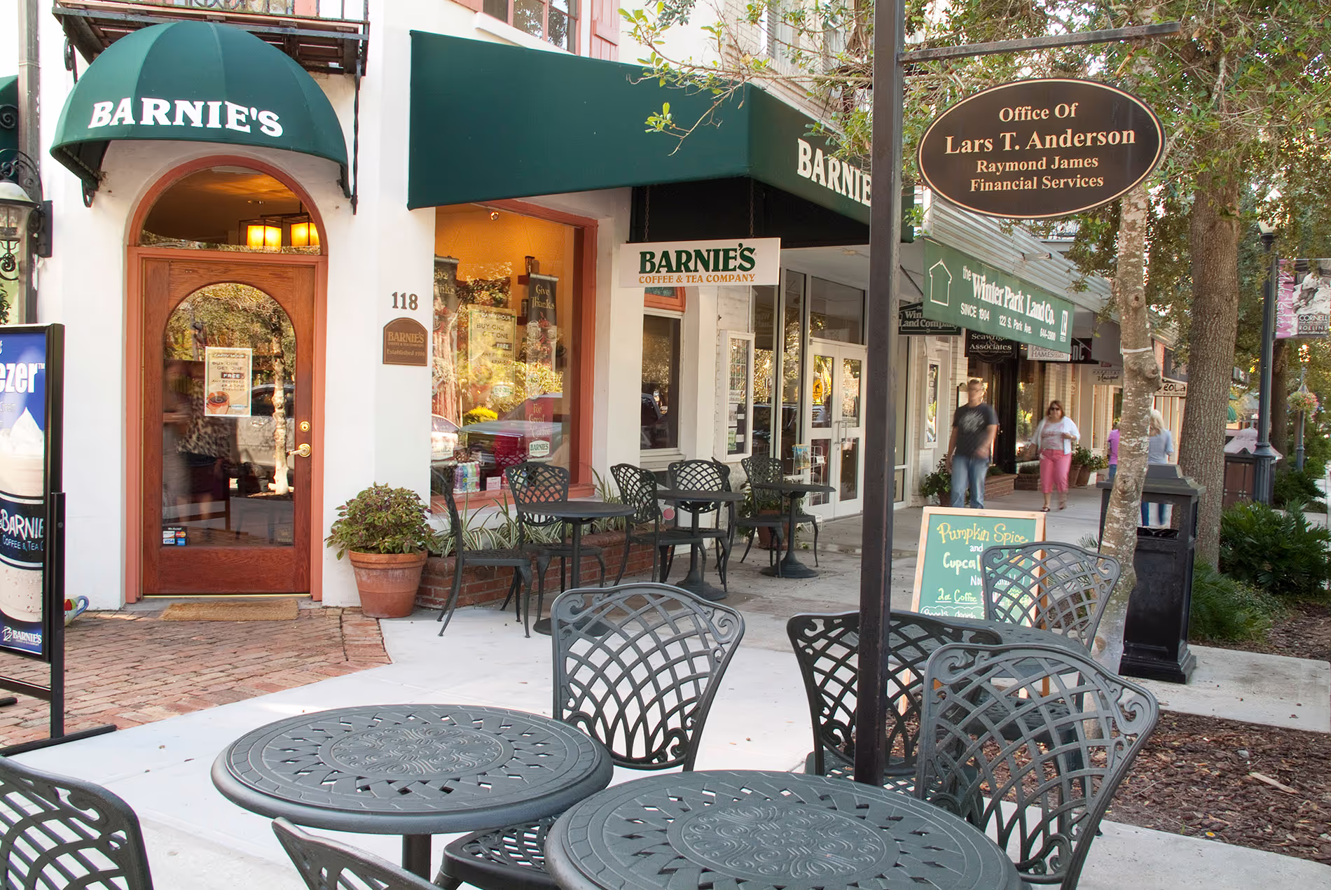 Outdoor dining scene in downtown Winter Park, Florida, with people enjoying meals under umbrellas along a charming, tree-lined street.