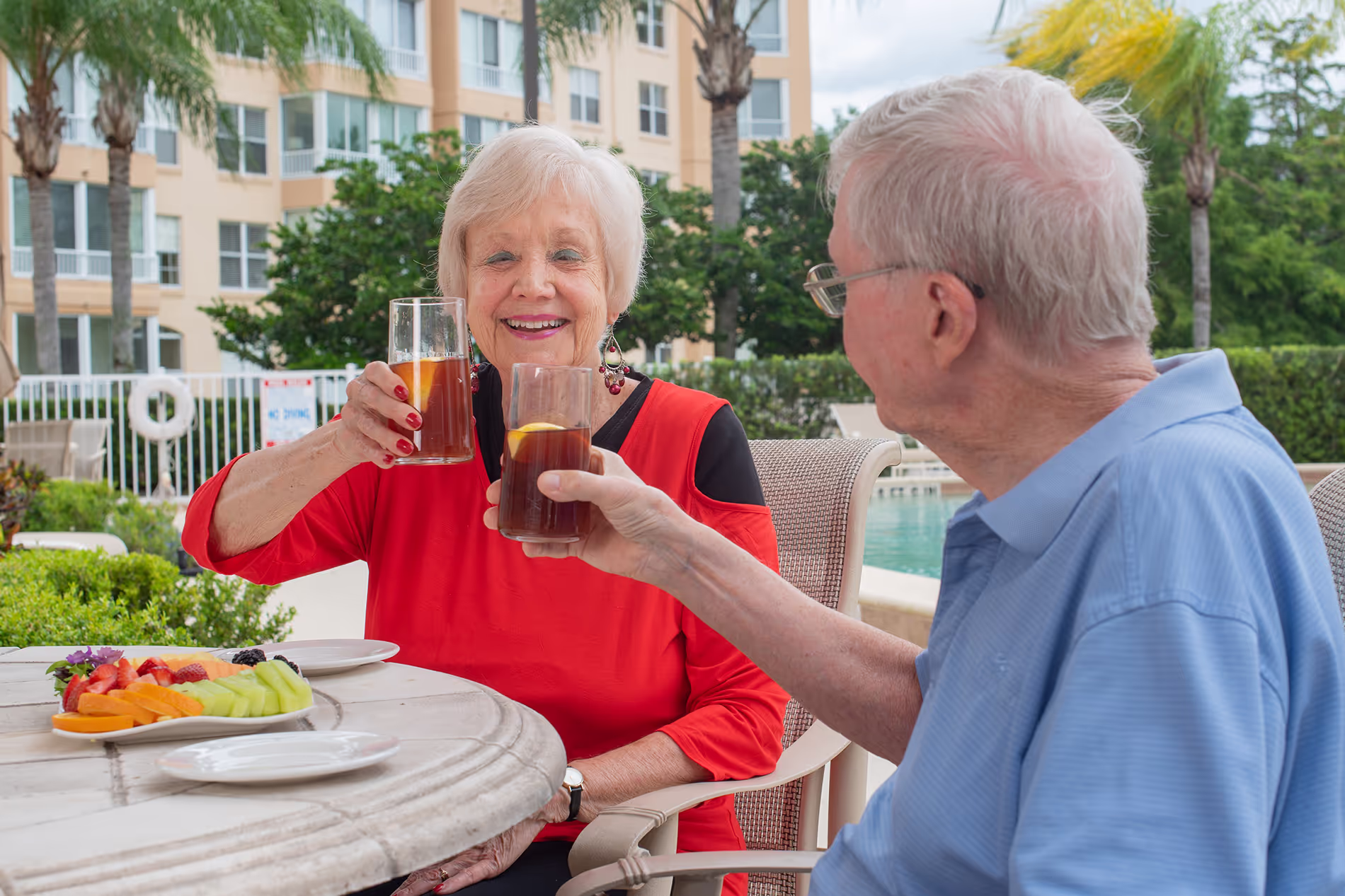 Couple relaxing by the pool, sipping iced tea and enjoying a plate of fresh fruit in a sunny, peaceful setting.