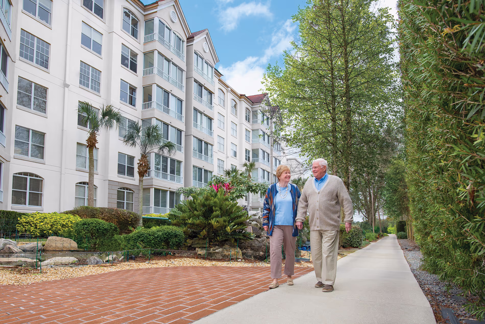 Older couple walking together in front of one of the apartment buildings on the retirement community campus, enjoying a peaceful and leisurely moment.
