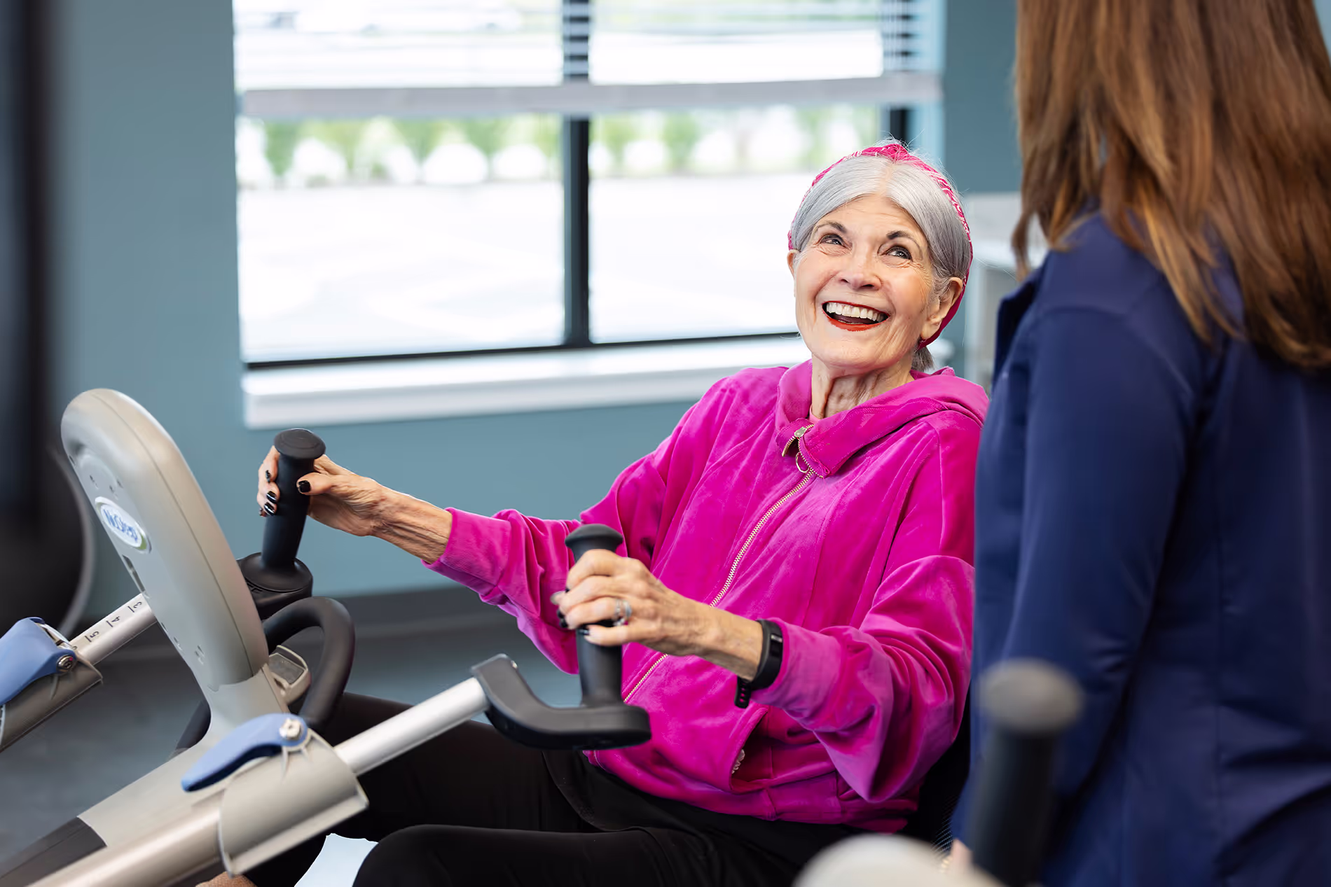 Older adult using a cycling exercise machine with guidance from a trainer, both focused and engaged in a supportive fitness session.