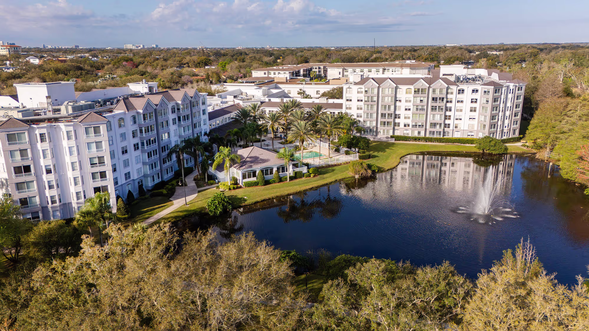 Aerial view of apartment buildings next to a serene lake, with surrounding greenery and walking paths enhancing the scenic setting.