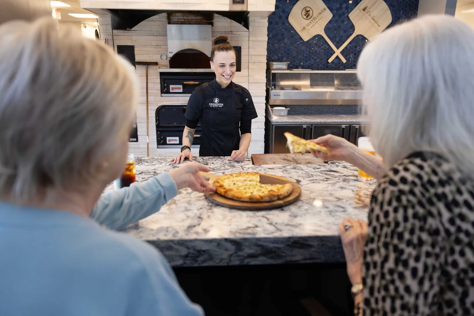 Chef serving a freshly made pizza to an older woman at the dinner table, with a warm and welcoming smile.