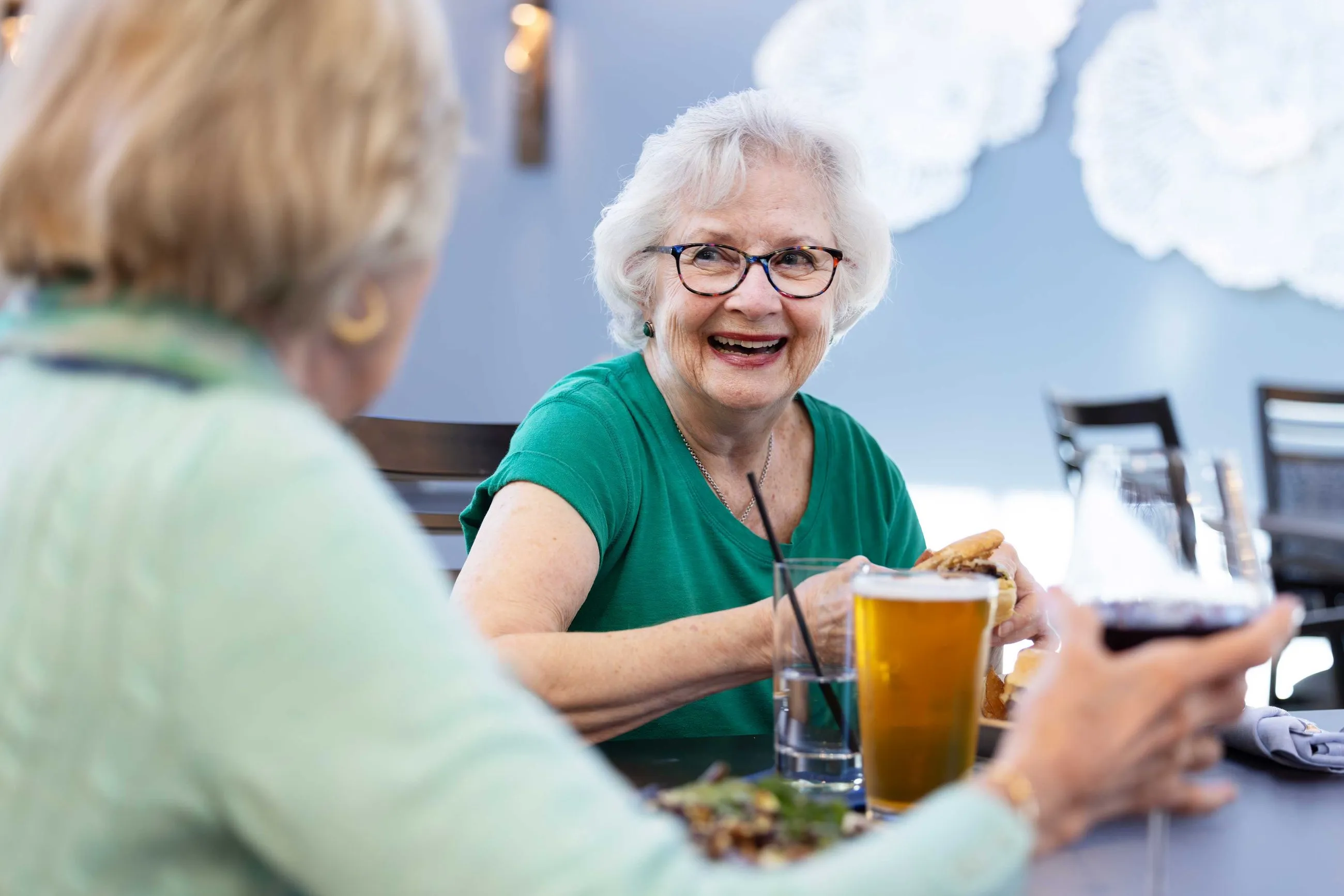 Older woman smiling and enjoying a meal with a beer, sitting with a friend in a relaxed, cheerful setting.