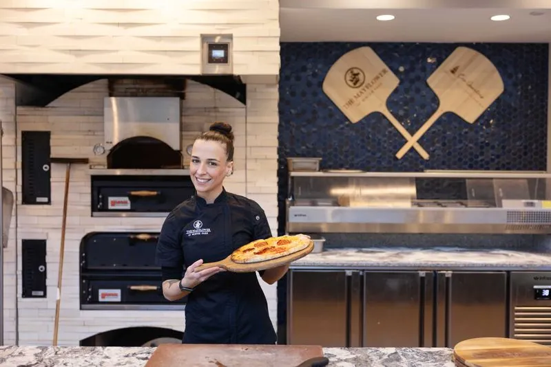 Chef proudly holding a freshly made pizza in front of a stone oven, showcasing their culinary creation.