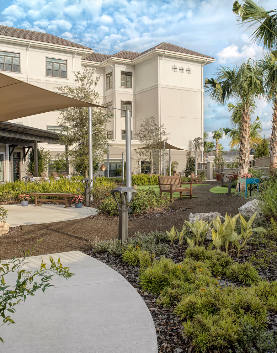 Peaceful memory care garden located behind a building, featuring lush greenery, flowers, and a tranquil atmosphere for reflection and relaxation.