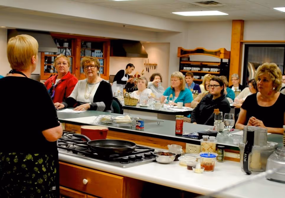 Woman teaching a cooking class with seated students watching and kitchen tools and ingredients on counters.