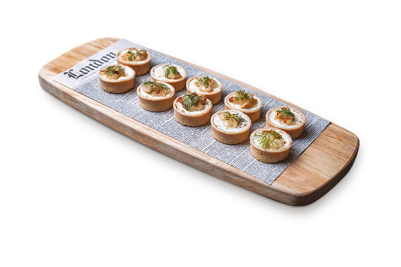 Wooden platter with ten small tart appetizers garnished with herbs on printed paper.