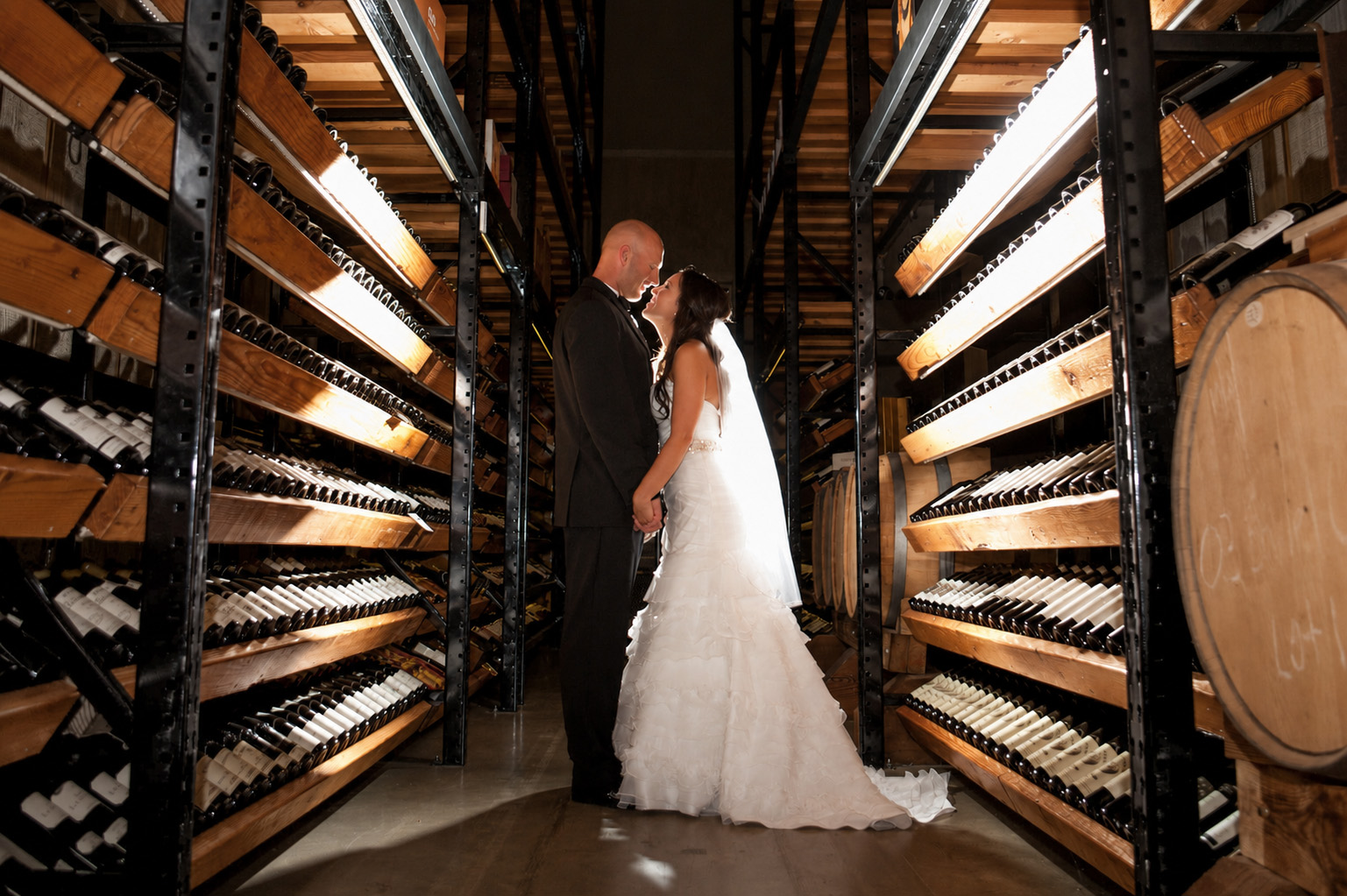Bride and groom holding hands and gazing at each other in a wine cellar aisle.