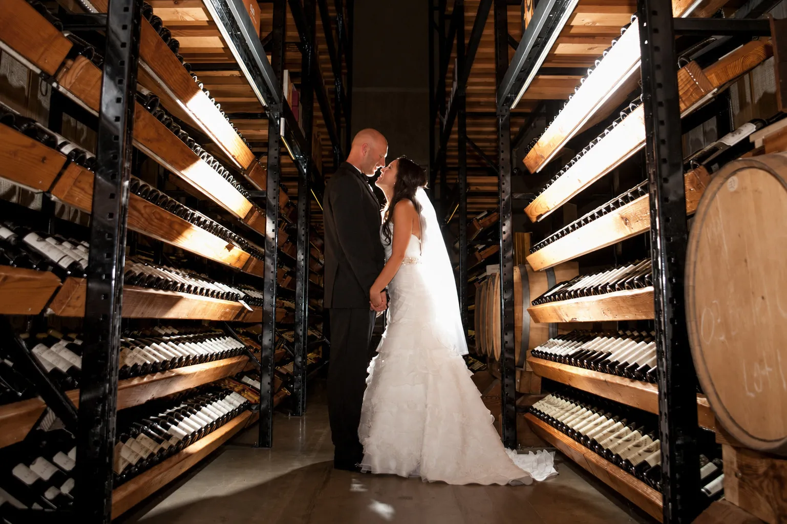Bride and groom holding hands and gazing at each other in a wine cellar aisle.