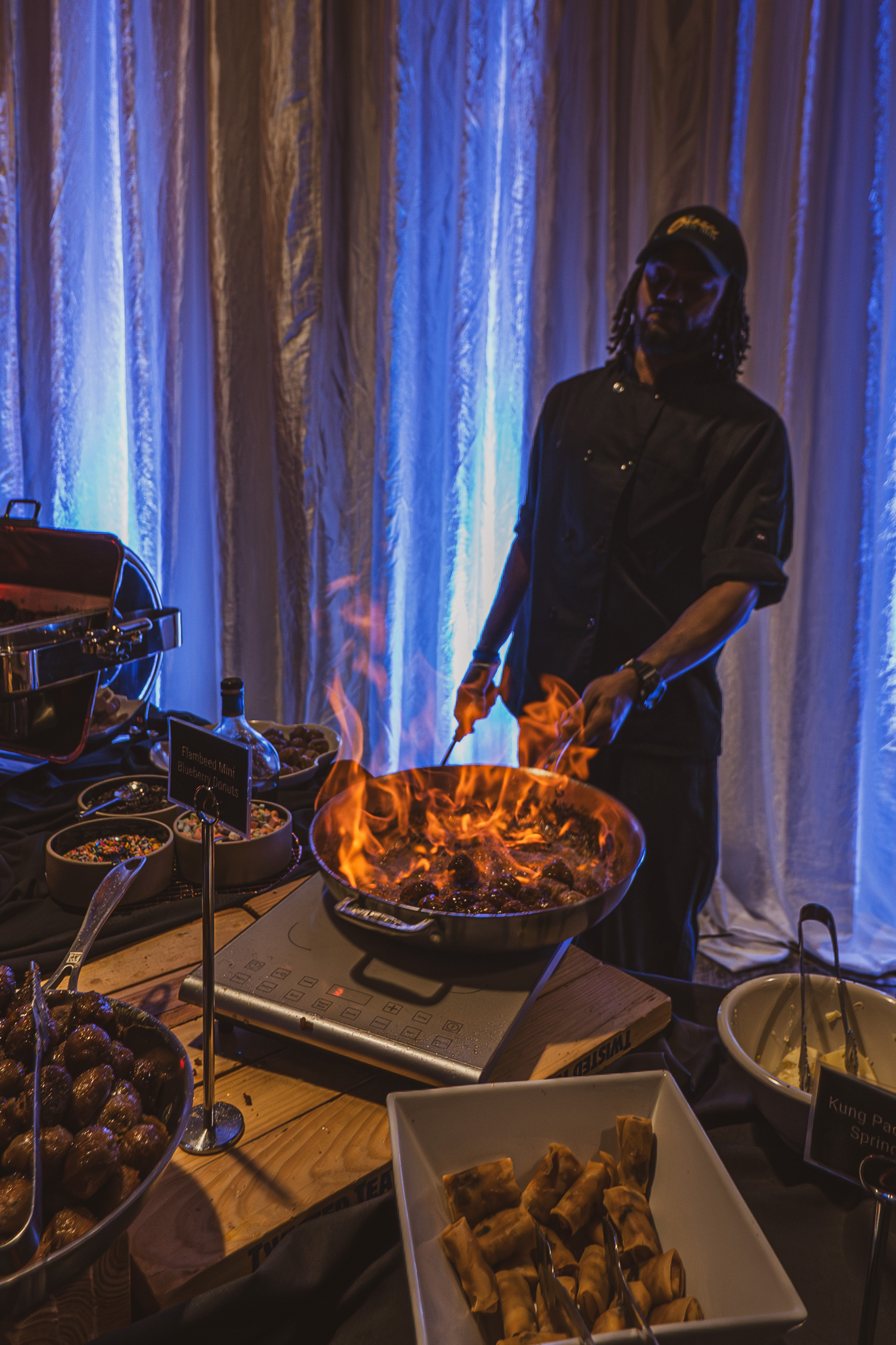 Chef cooking flaming dish on a stove at a buffet with appetizers and blue-lit curtains behind.