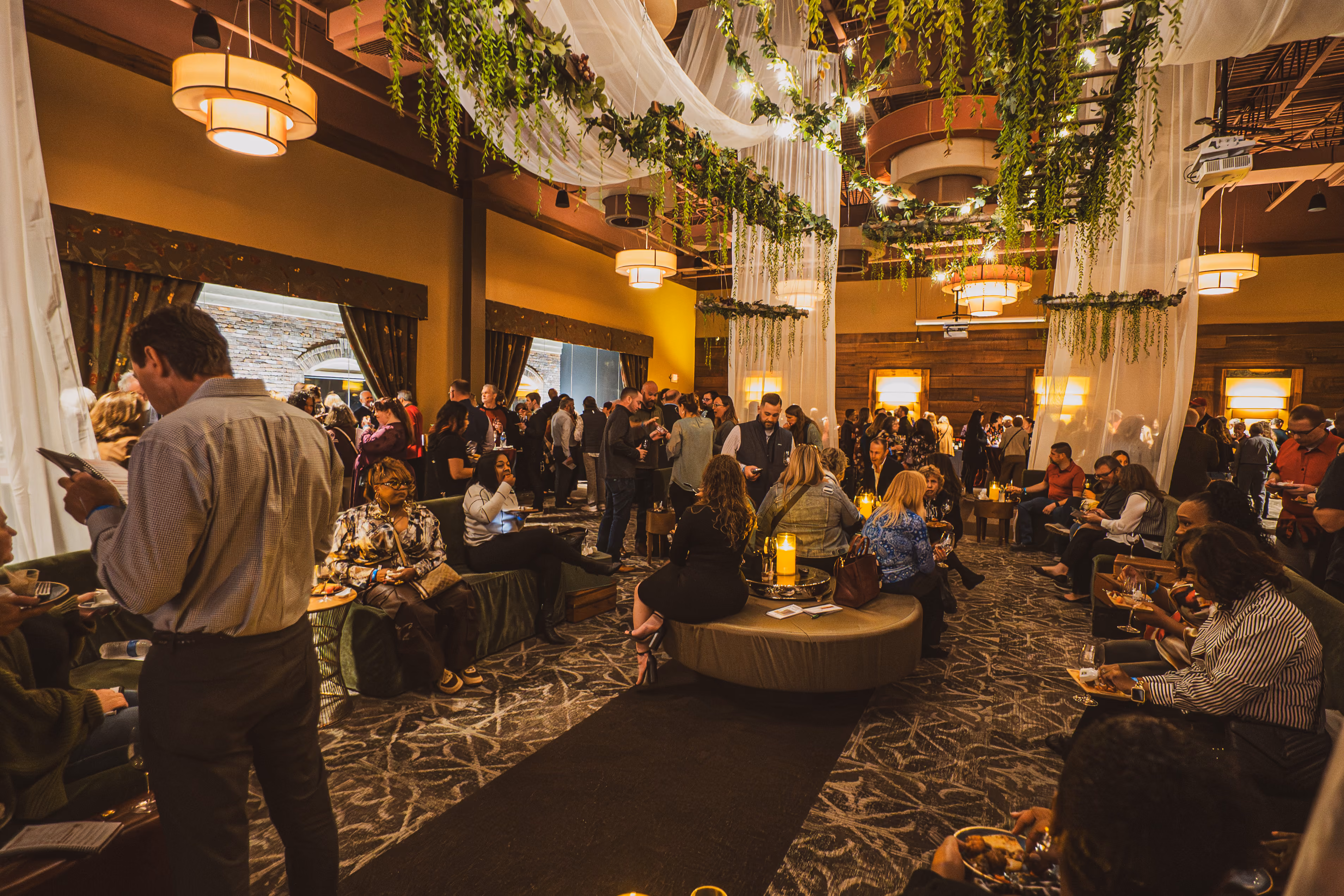 Crowded indoor event with people eating, socializing under hanging greenery and warm lighting.