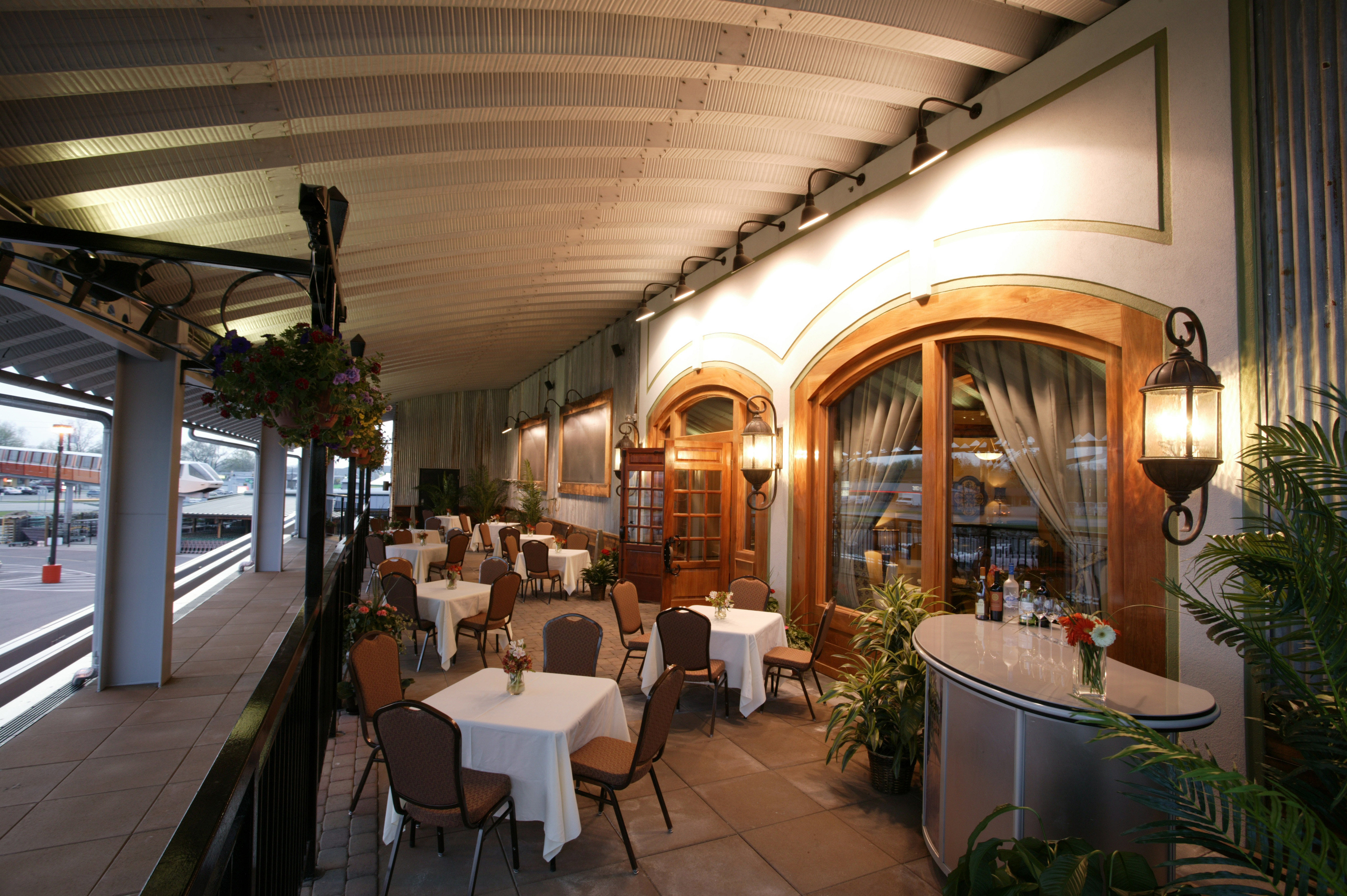 Outdoor covered restaurant patio with tables, chairs, plants, and warm wall lamps in the evening.
