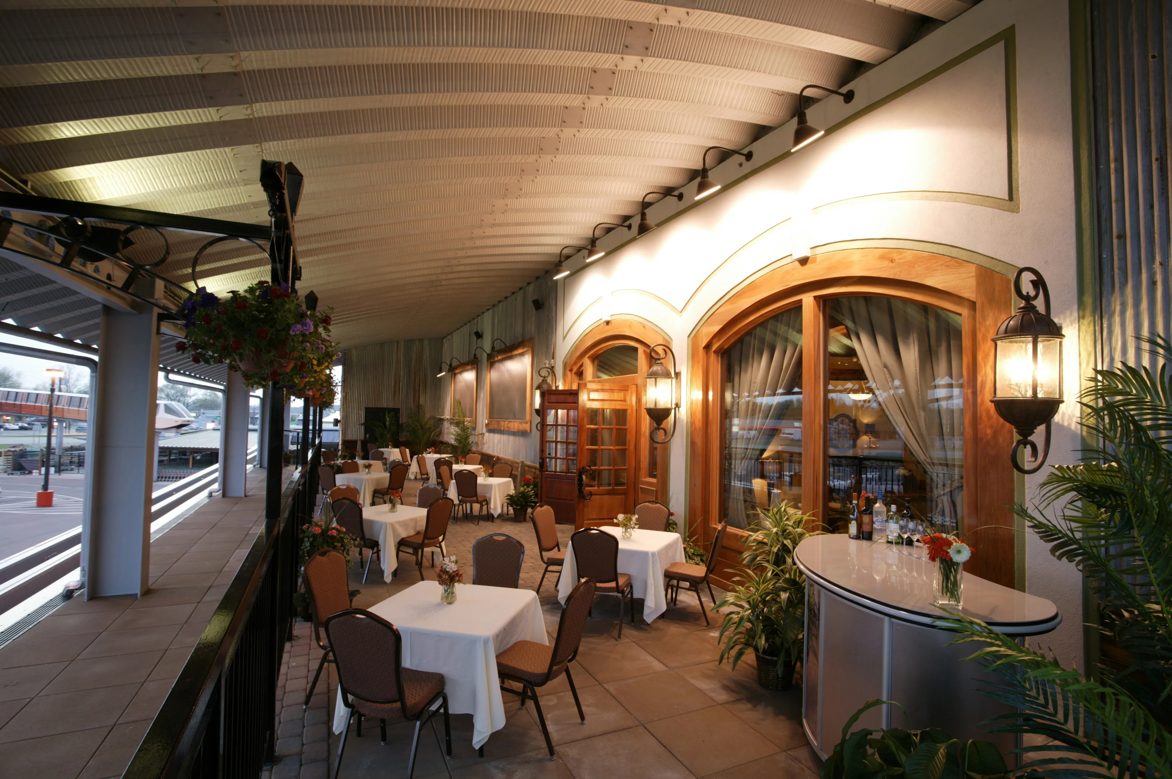 Outdoor covered restaurant patio with tables, chairs, plants, and warm wall lamps in the evening.
