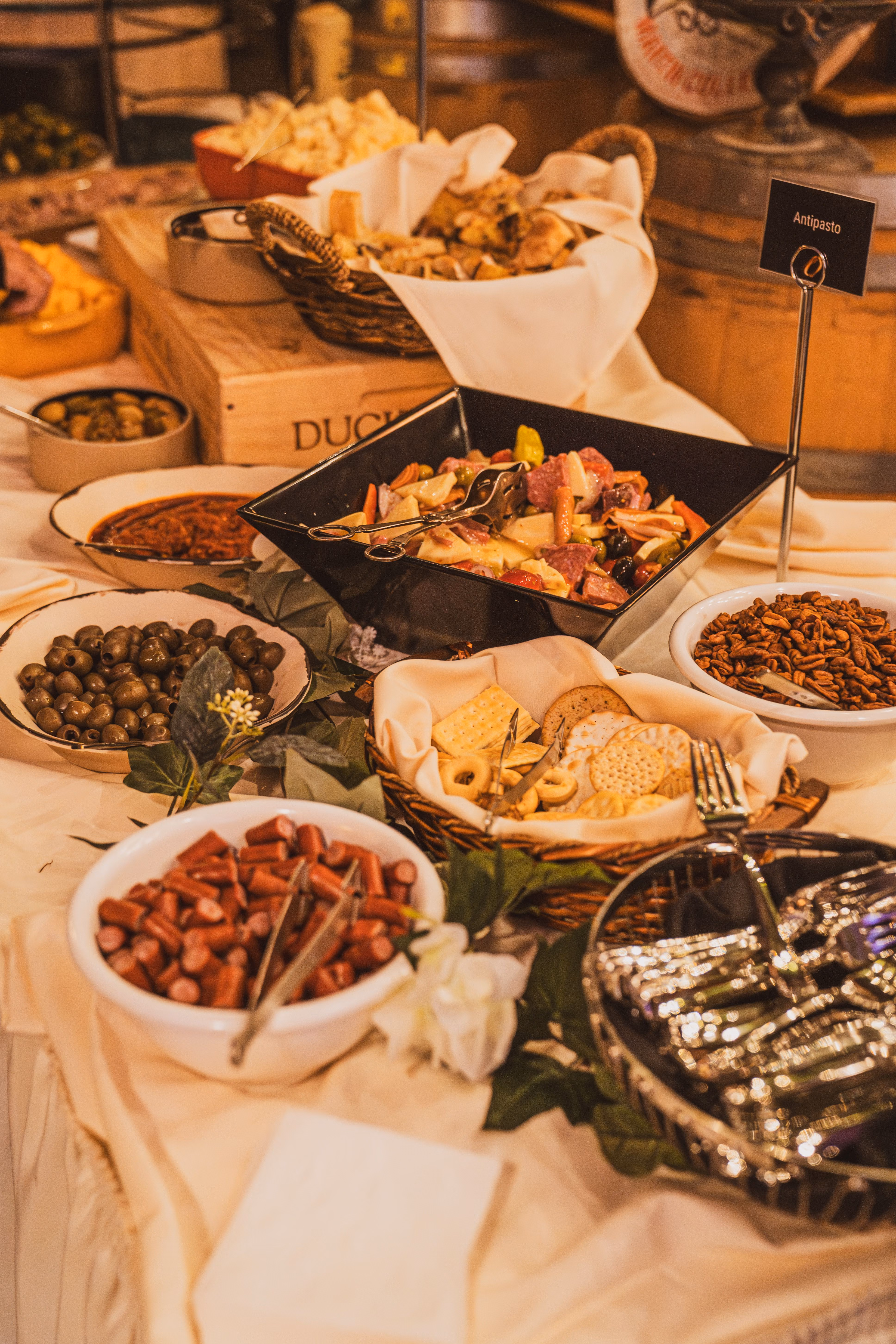 Buffet table with antipasto, olives, crackers, sausage pieces, nuts, and silverware.