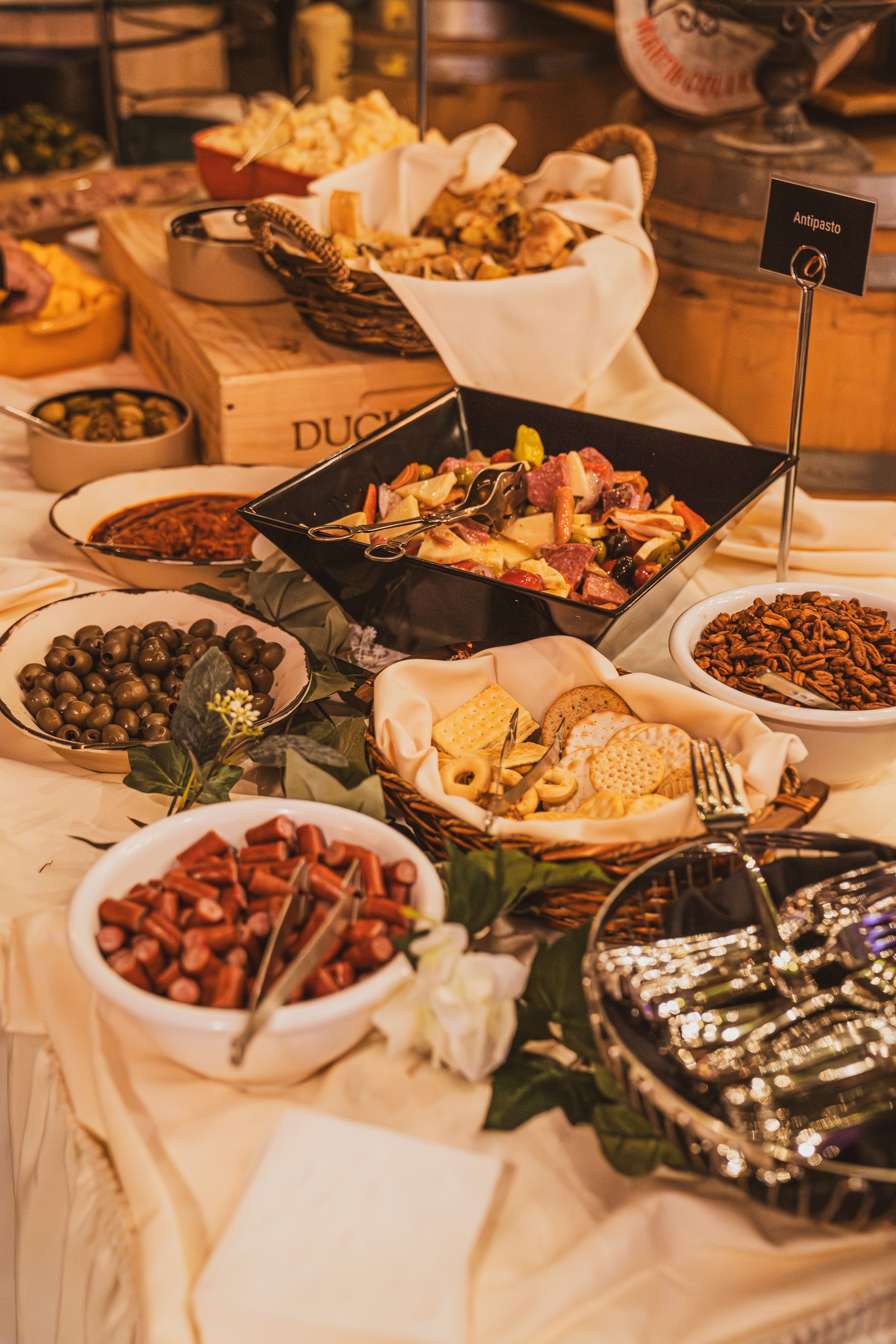 Buffet table with antipasto, olives, crackers, sausage pieces, nuts, and silverware.