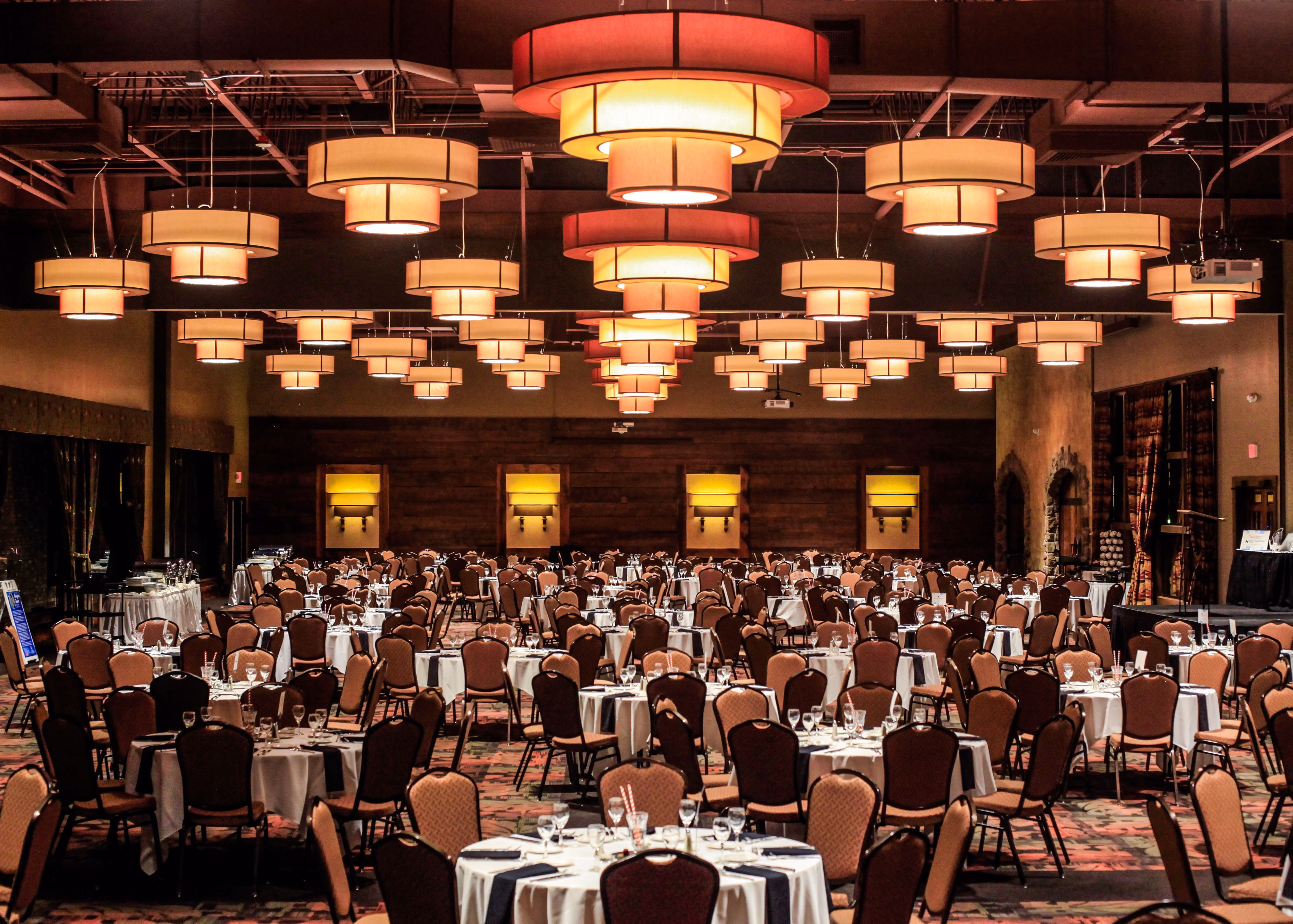 Large banquet hall with round tables and chairs, set with glassware and cutlery under hanging lights.