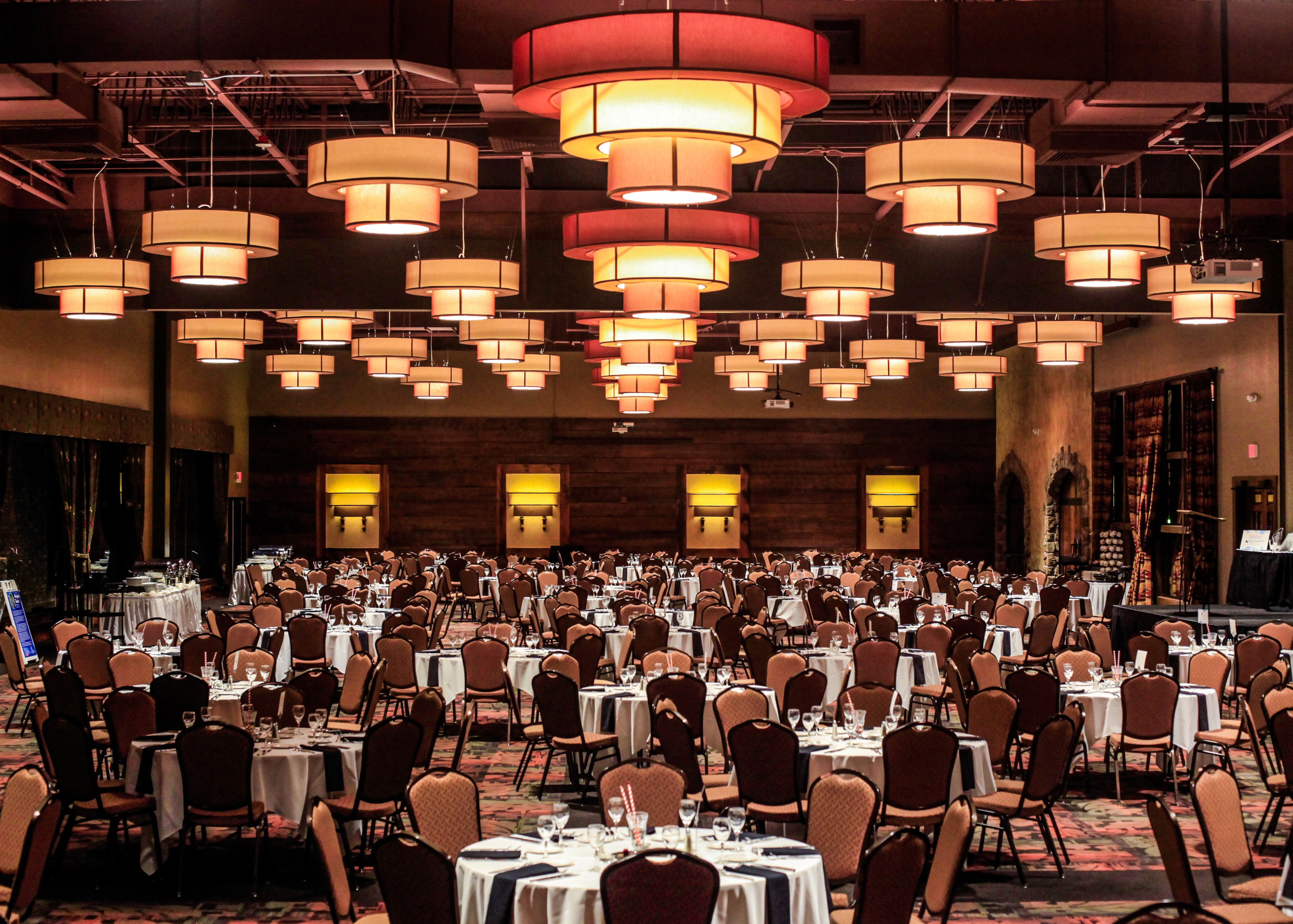 Large banquet hall with round tables and chairs, set with glassware and cutlery under hanging lights.