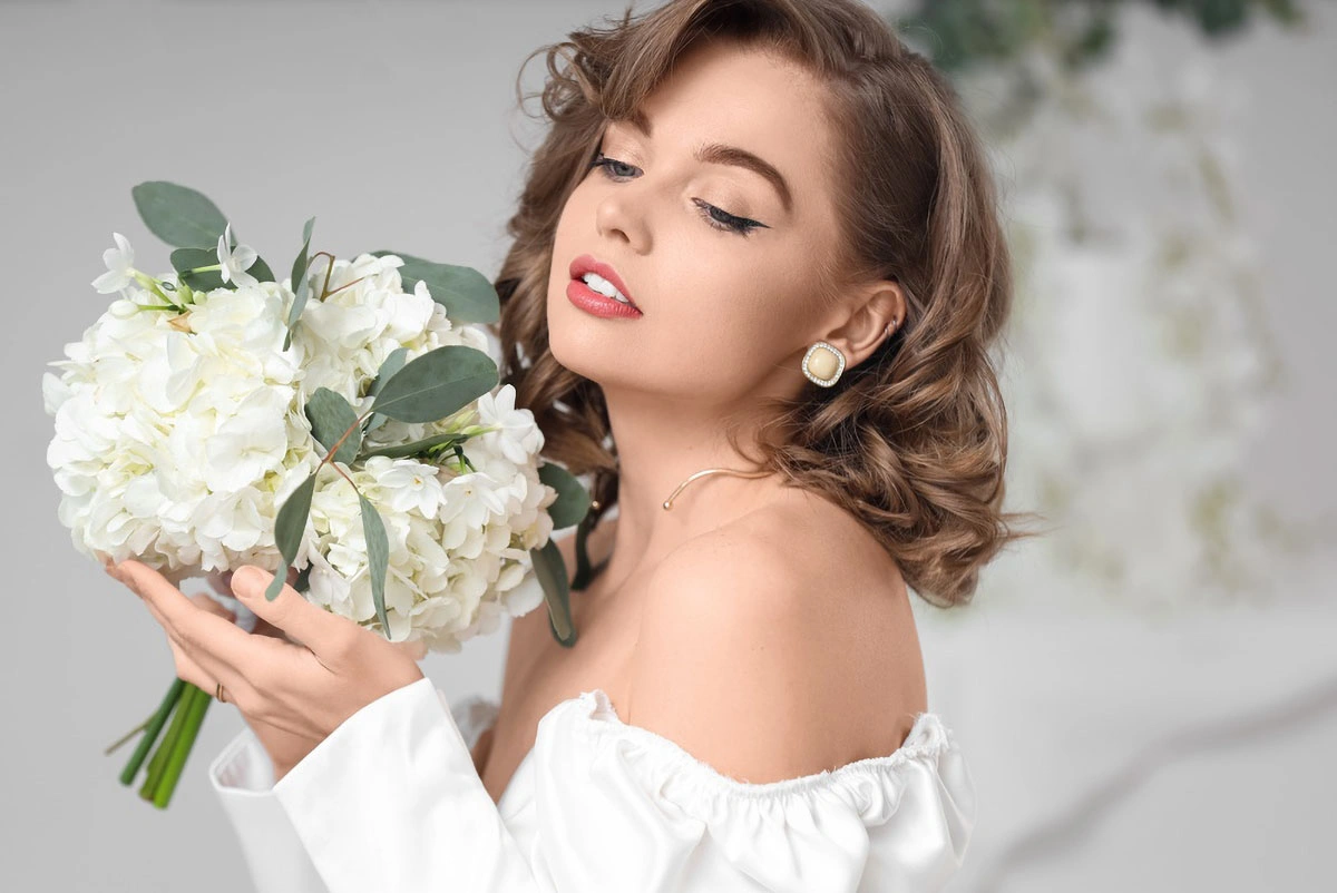 Woman with curled brown hair holding a bouquet of white flowers while wearing pearl earrings and an off-shoulder white blouse.