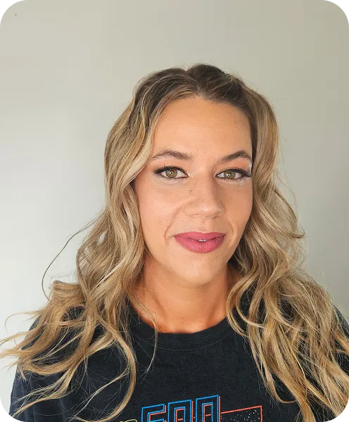 Smiling woman with long wavy blonde hair and pink lipstick wearing a black shirt standing against a light gray background.