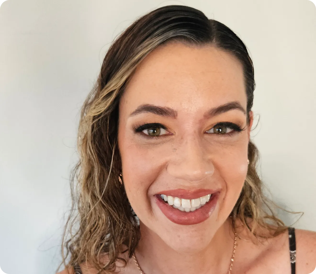 Close-up of a smiling woman with curly highlighted hair, wearing makeup and a necklace.