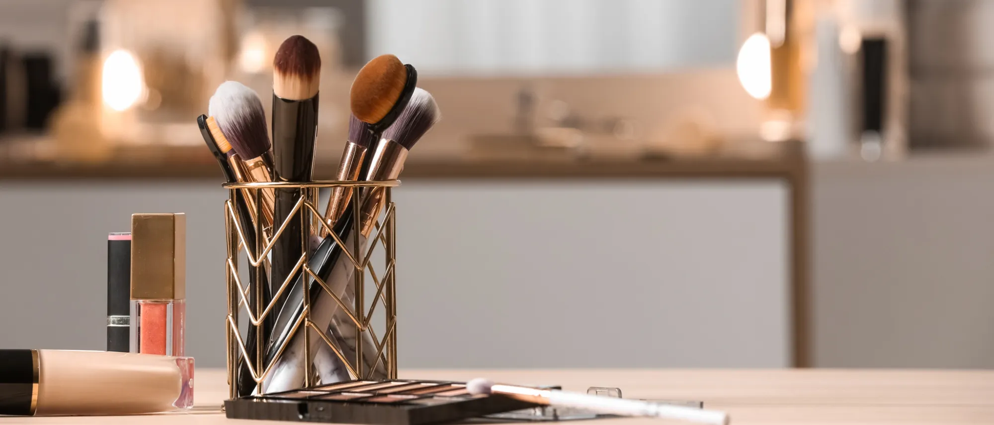 Set of makeup brushes in a gold holder alongside lip gloss and a makeup palette on a wooden surface.