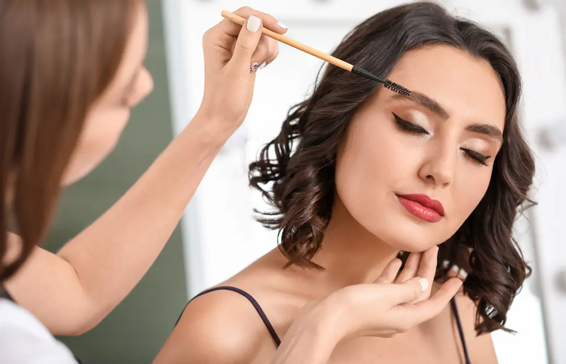 Makeup artist grooming eyebrows and applying glam makeup to female client