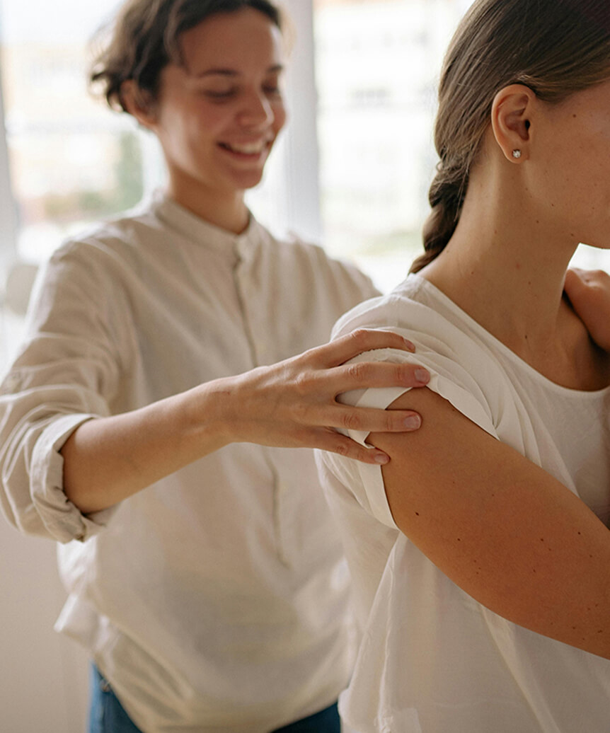 Woman in white shirt smiling while touching another woman's shoulder indoors near window light.