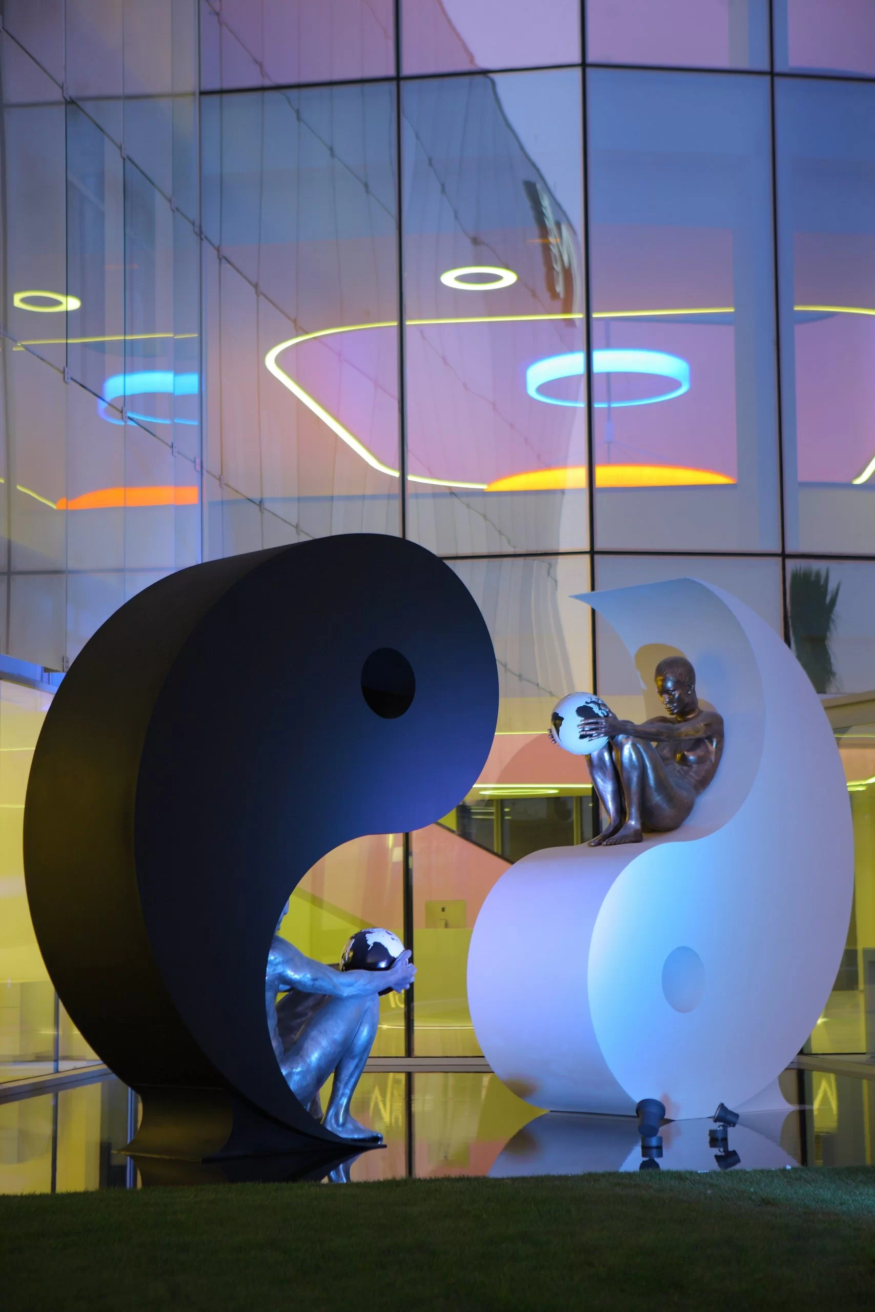 Escultura de yin y yang con figuras humanas metálicas, en la entrada de IM Gournay, sosteniendo globos terráqueos frente a edificio moderno.