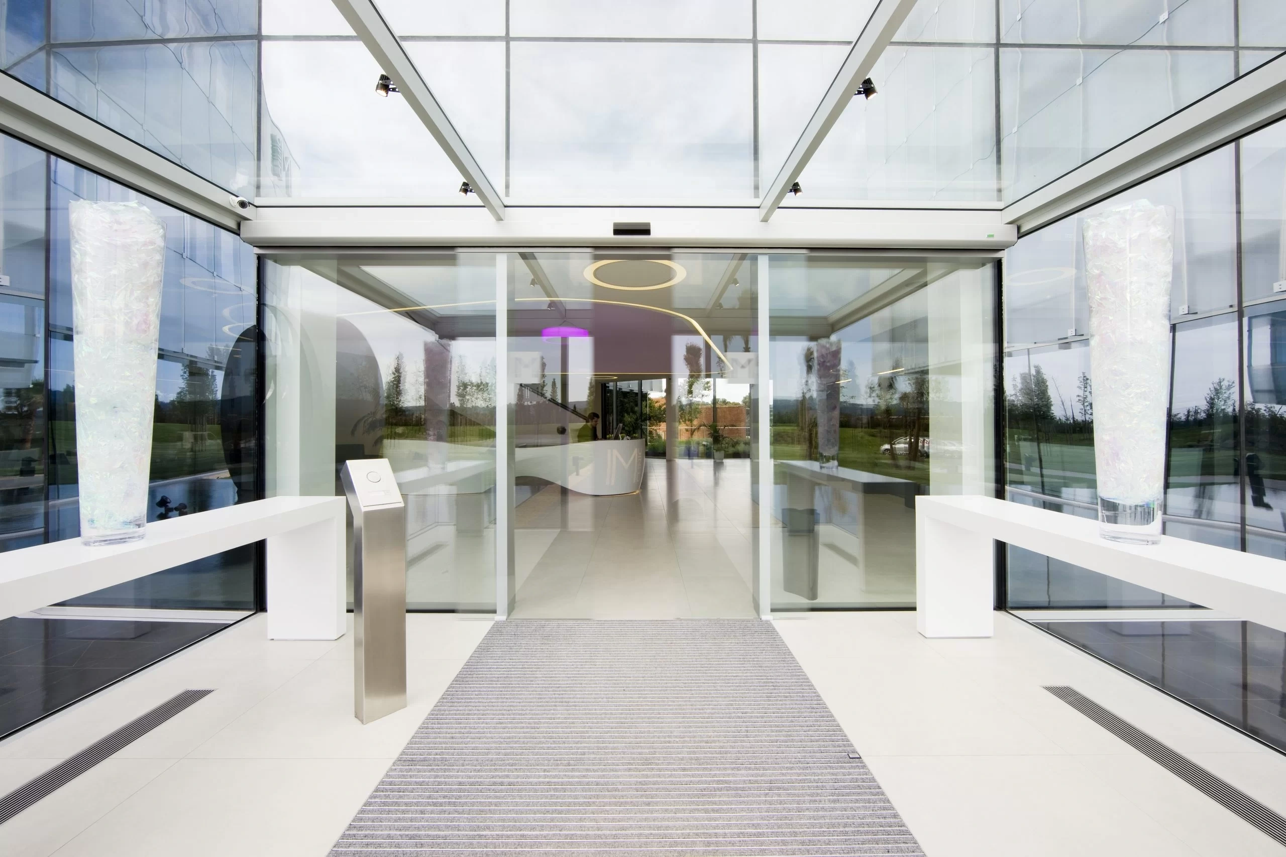 Entrada moderna de IM Gournay con puertas de cristal automáticas, piso blanco y grandes jarrones decorativos.