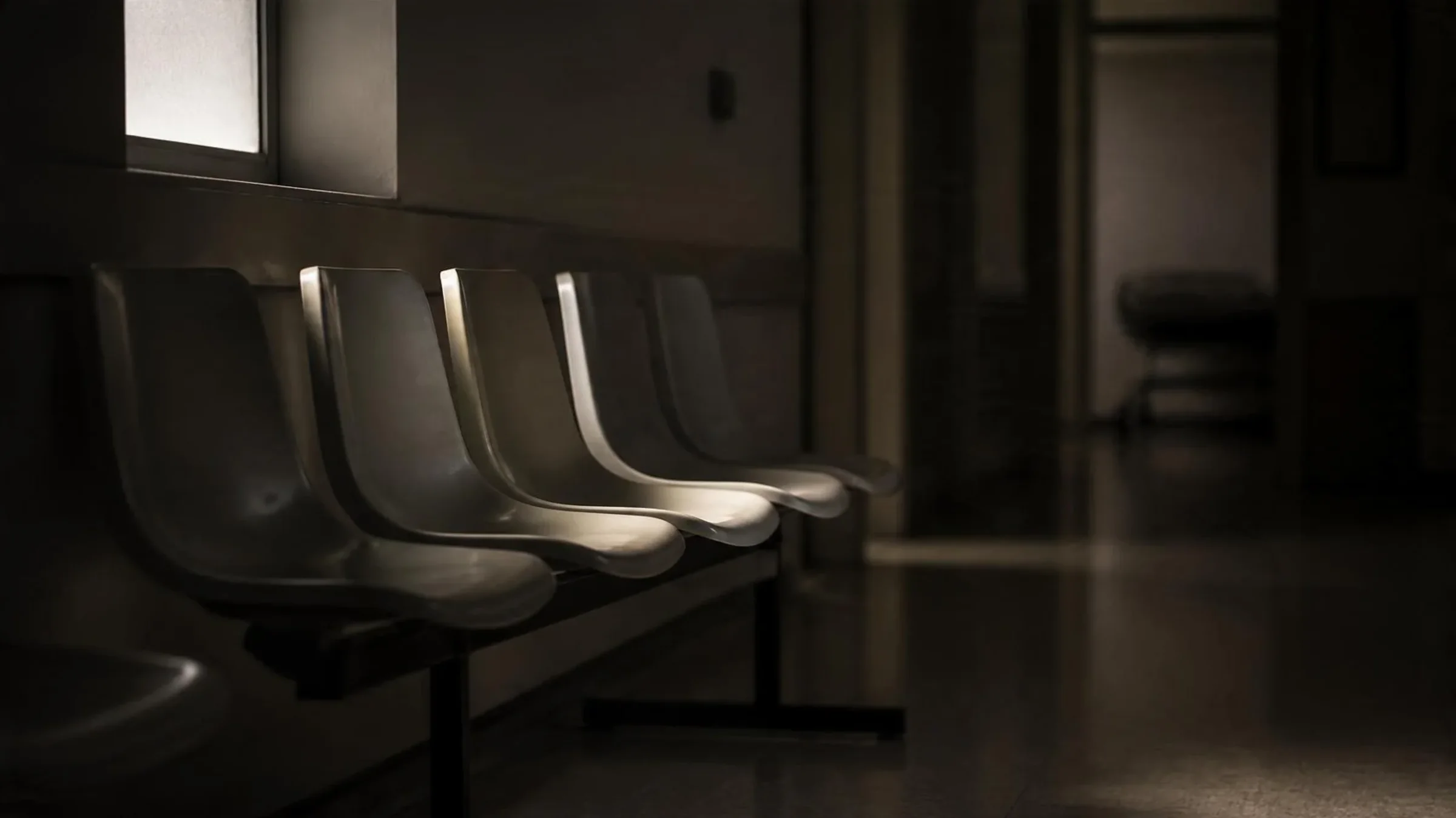 Empty waiting-room chairs in a dim hospital corridor, lit by soft side light, evoking NHS waiting lists for FFS