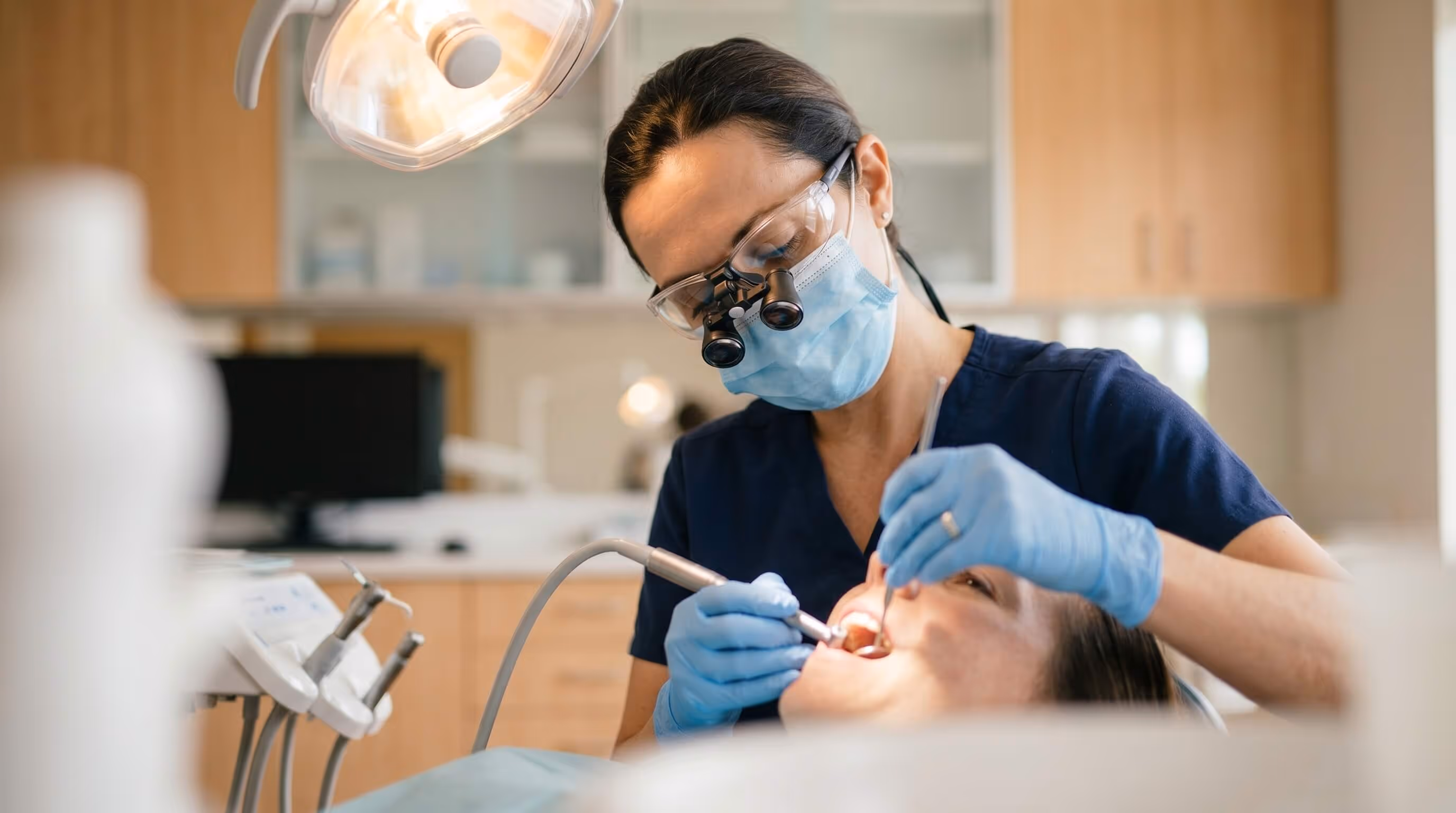 Dentist wearing magnifying glasses and mask performing dental procedure on a patient in a clinic.
