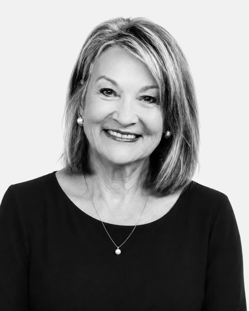 Smiling older woman with shoulder-length hair wearing a black top, pearl earrings, and necklace.