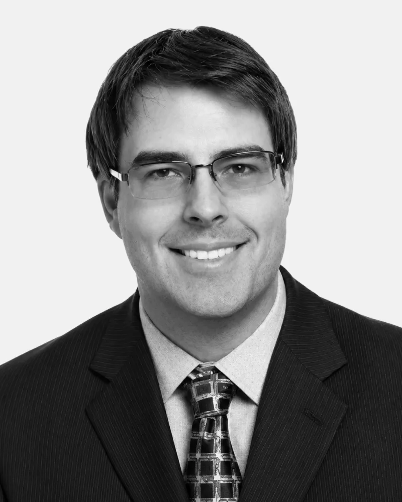 Smiling man with glasses wearing a suit, dress shirt, and patterned tie against a plain background.