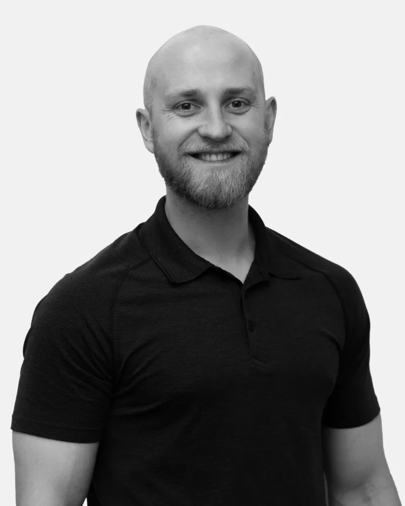 Smiling bald man with a beard wearing a black polo shirt against a plain background.