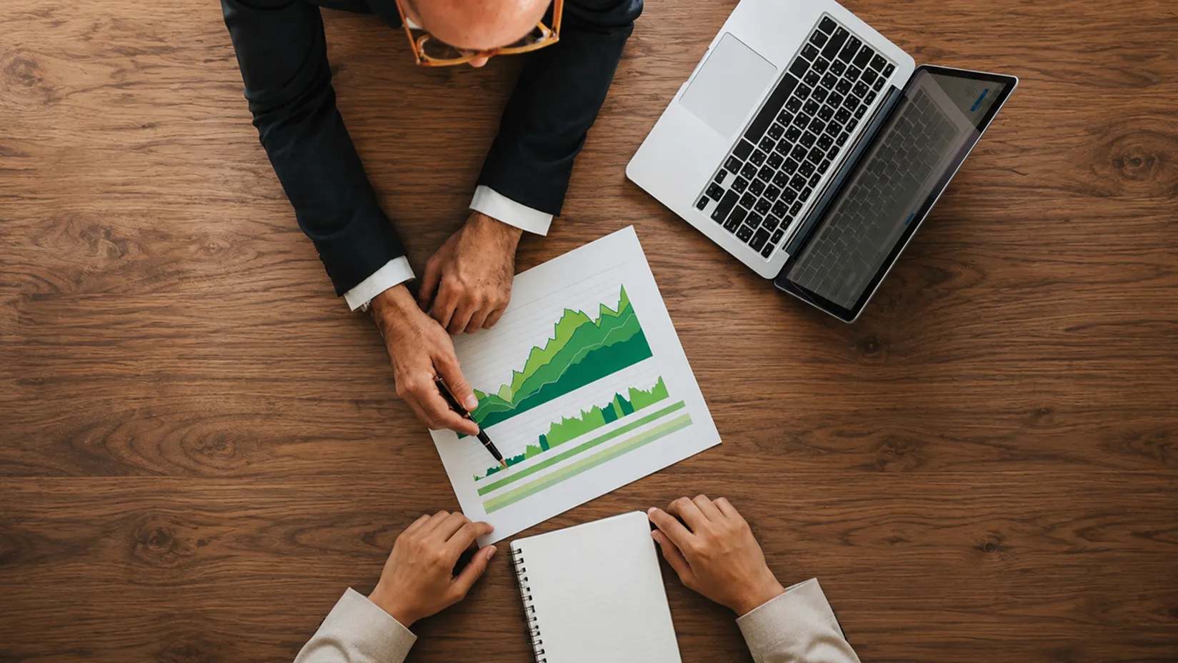 Two people analyzing green growth charts with laptop and notebook on a wooden table.