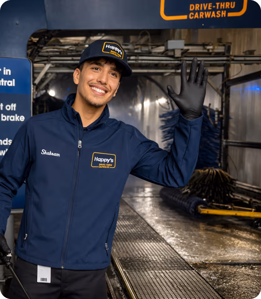 Smiling car wash employee wearing a navy uniform and black gloves waving inside a drive-thru carwash.