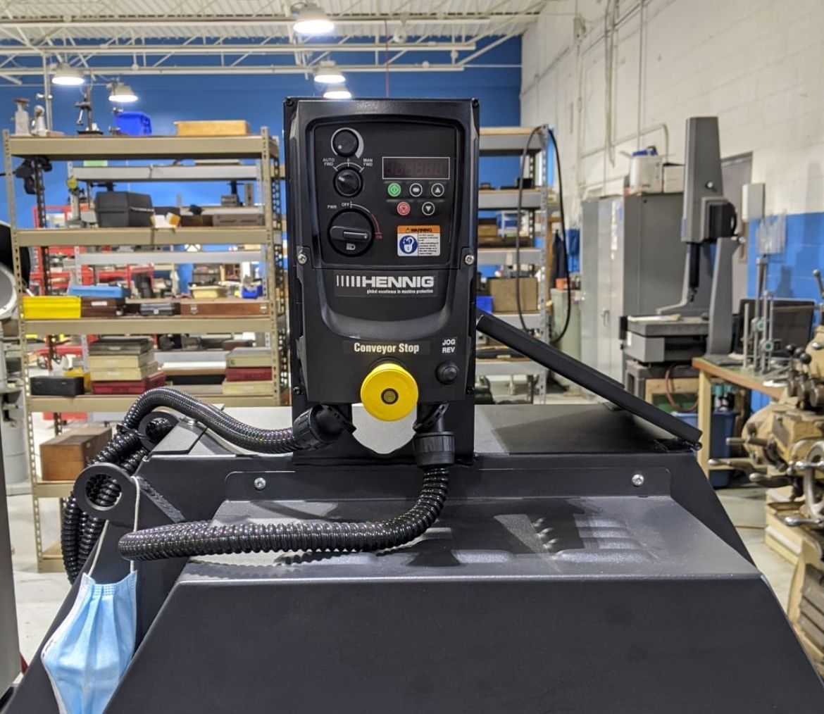 Close-up of a Hennig conveyor stop control panel in an industrial workshop with shelves and machinery in the background.